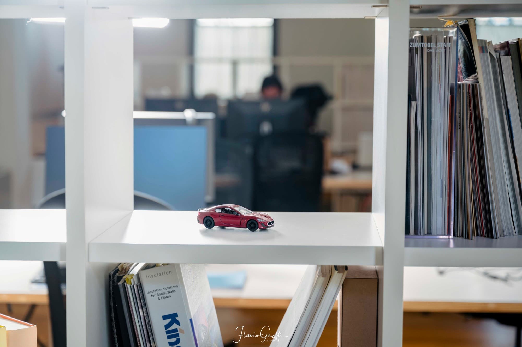A model Maserati on a bookcase, with the office and its screens blurred in the background